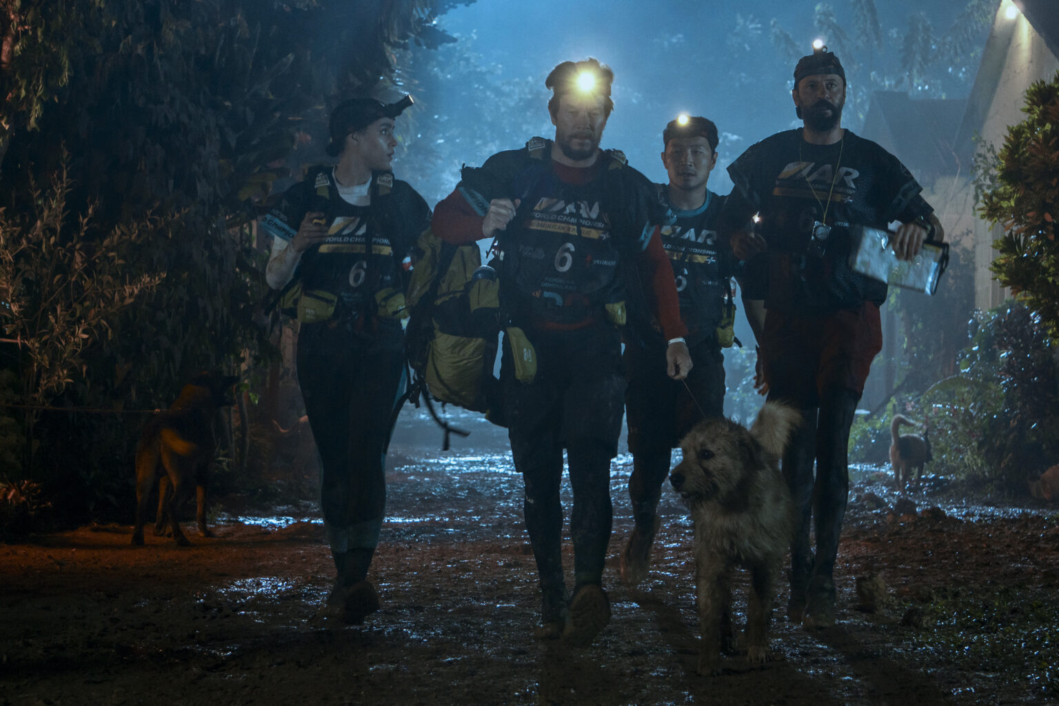 Cztery osoby noszące czołówki i sprzęt do wyścigów przygodowych idą razem błotnistą ścieżką nocą, w towarzystwie kudłatego psa. Scena jest oświetlona czołówkami i niebieskim światłem.