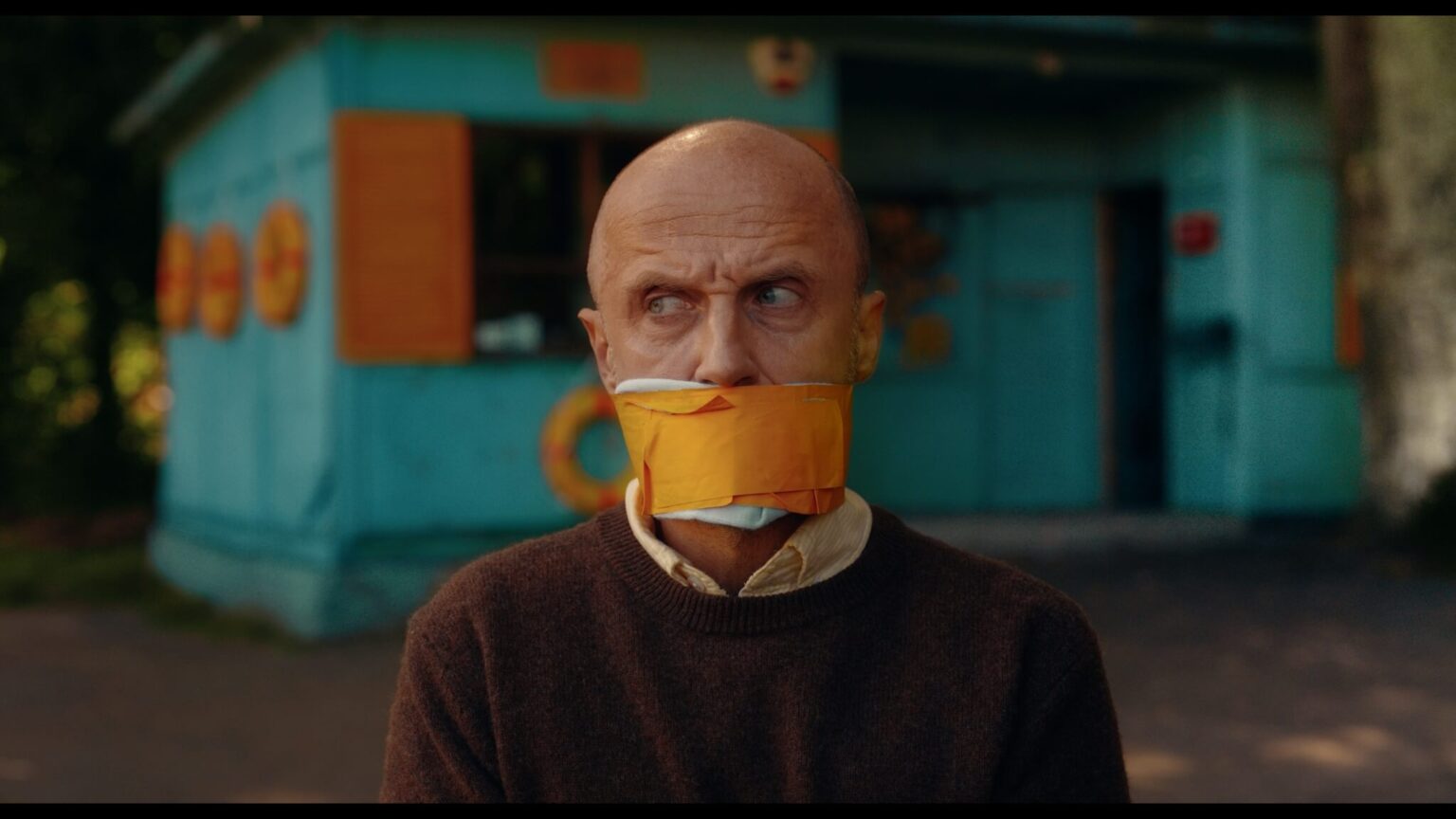 Łysy mężczyzna w brązowym swetrze siedzi na zewnątrz z ustami zakrytymi maską i kawałkiem pomarańczowej taśmy. Patrzy w bok z zagadkowym wyrazem twarzy, przed niebieskim budynkiem z pomarańczowymi okiennicami.