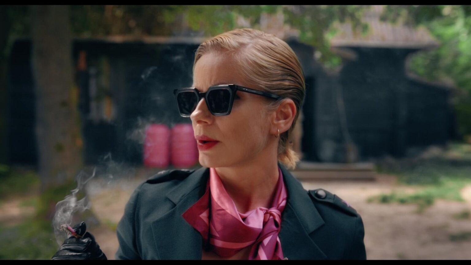 Kobieta z krótkimi, zaczesanymi do tyłu blond włosami i czarnymi okularami przeciwsłonecznymi trzyma papierosa, dym unosi się w górę. Ma na sobie ciemny płaszcz i różowy szalik, stoi na zewnątrz przed rozmazanym budynkiem.