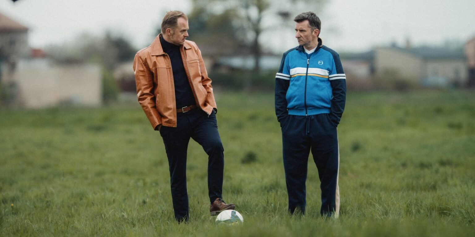 Dwóch mężczyzn stoi na trawiastym polu z piłką nożną u stóp. Jeden nosi brązową skórzaną kurtkę, czarne spodnie i golf, a drugi niebieski dres. Obaj wydają się prowadzić poważną rozmowę.