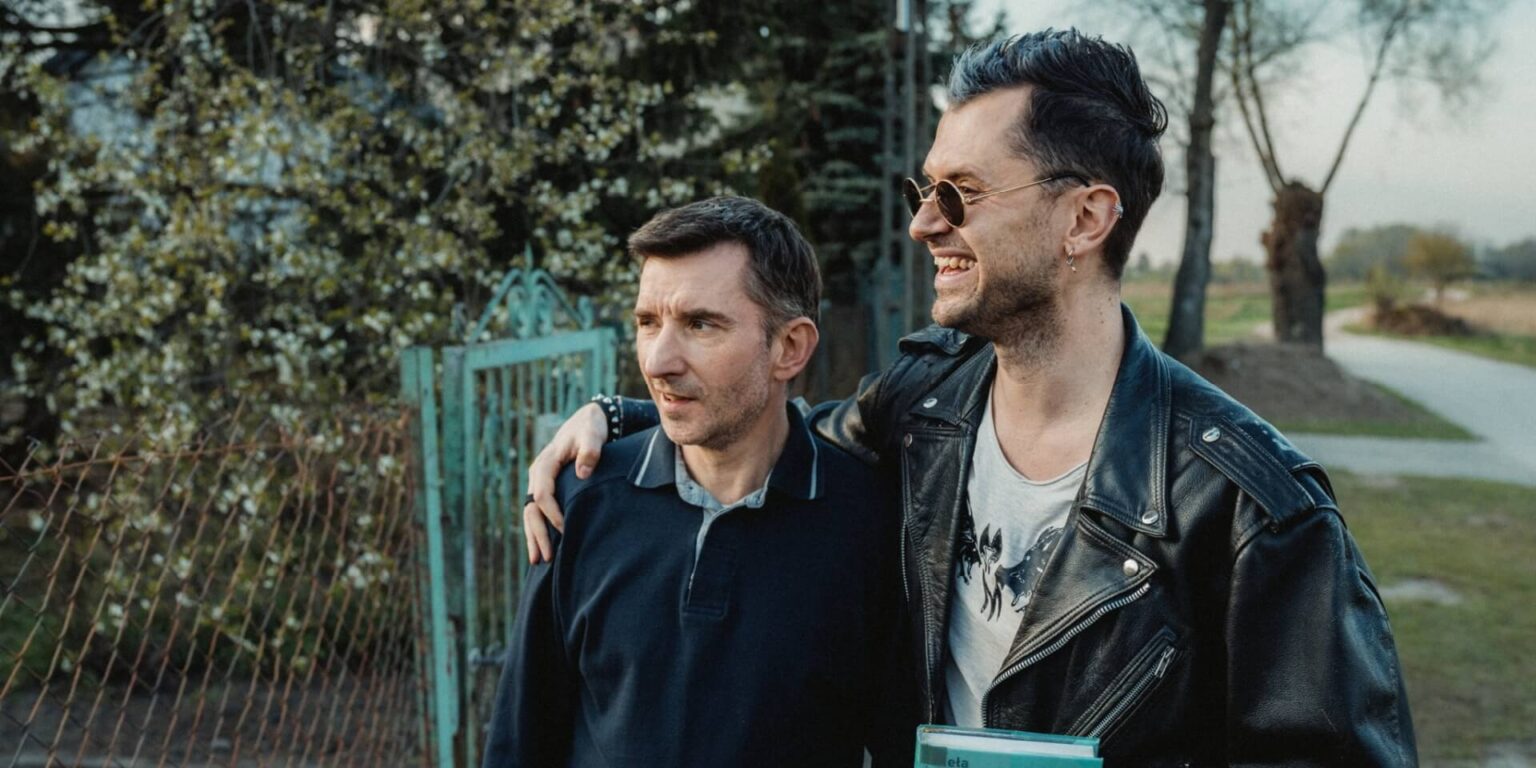 Dwóch mężczyzn stoi na zewnątrz przy płocie i drzewach. Jeden z nich, ubrany w ciemną koszulkę polo, patrzy przed siebie, podczas gdy drugi, w skórzanej kurtce i okularach przeciwsłonecznych, uśmiecha się i obejmuje ramieniem pierwszego z mężczyzn.