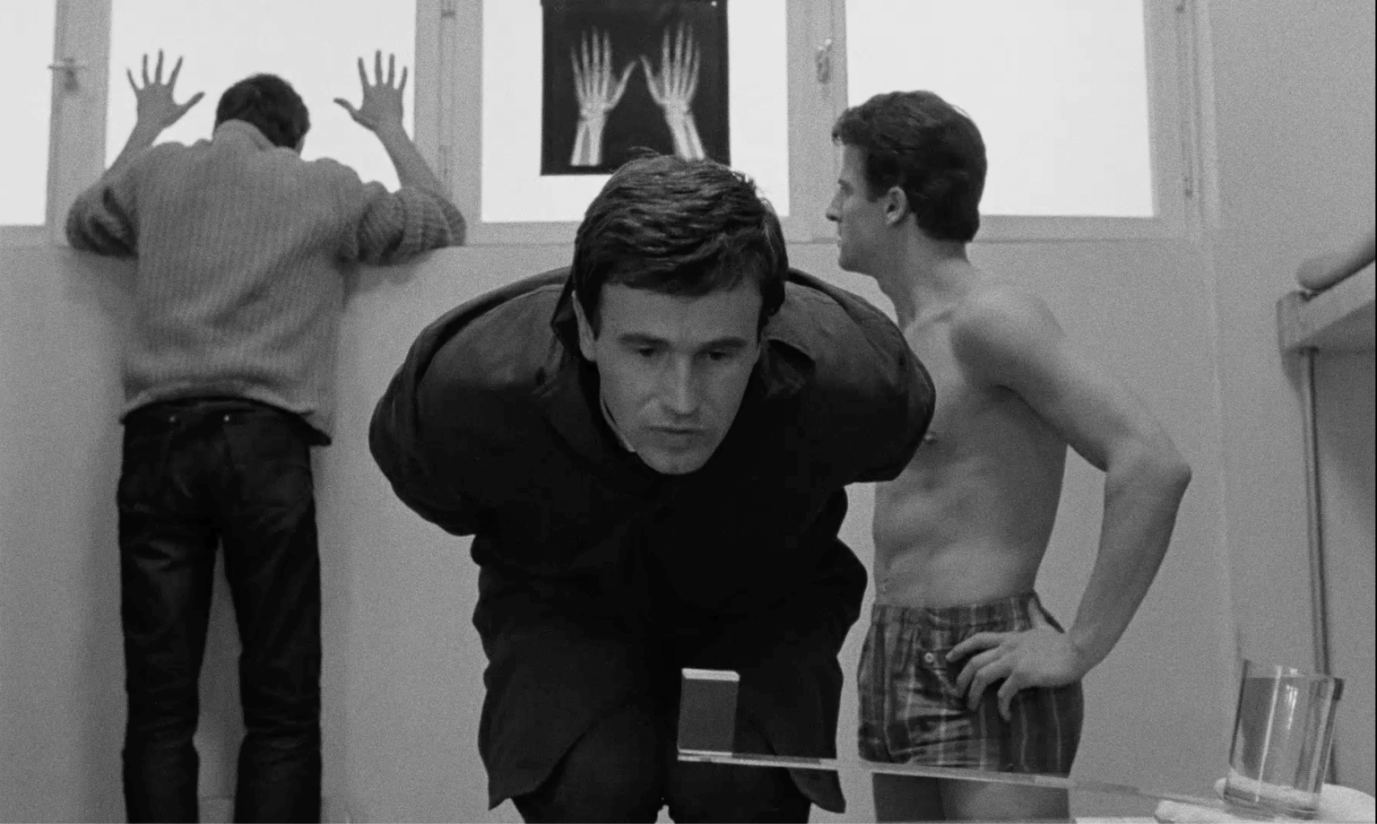 Trzech mężczyzn znajduje się w pokoju z białymi ścianami i wysokimi oknami. Jeden z nich stoi twarzą do okna, przyciskając dłonie do szyby, drugi pochyla się do przodu i patrzy w dół, a trzeci stoi bez koszuli w szortach w kratę. Na oknie znajduje się zdjęcie rentgenowskie dłoni.