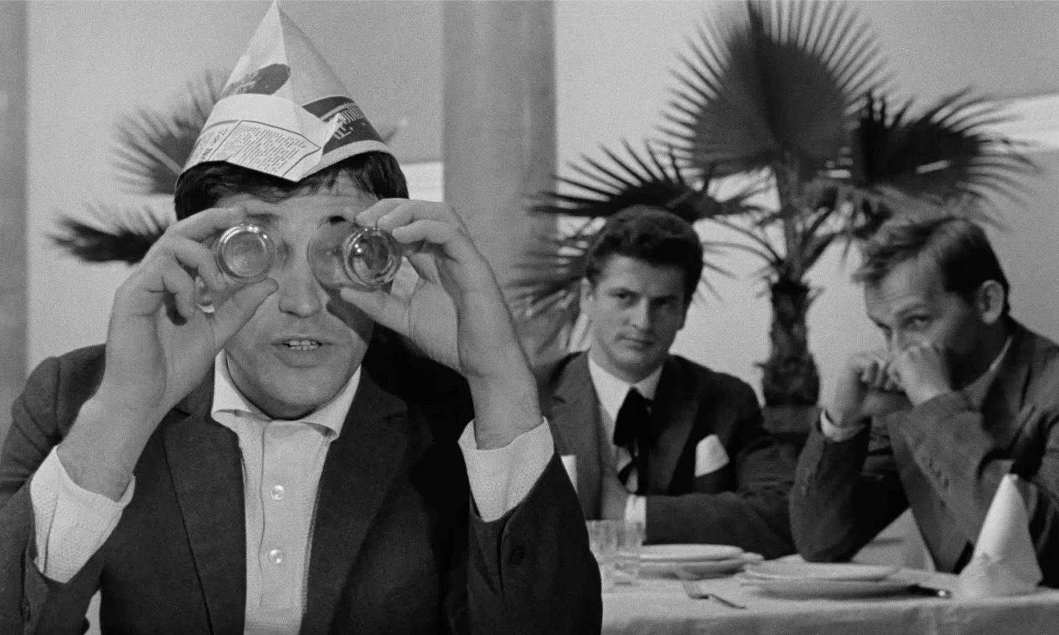Mężczyzna w papierowym kapeluszu trzyma dwie szklanki przed oczami jak lornetki, podczas gdy dwóch innych mężczyzn w garniturach siedzi przy stoliku za nim. Sceneria wydaje się być restauracją z doniczkowymi palmami w tle.