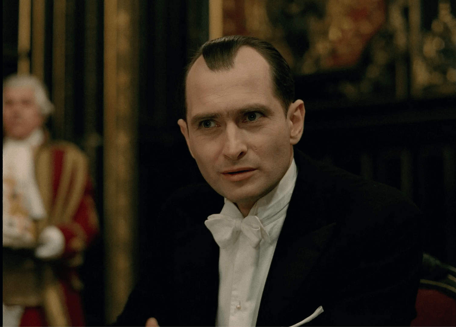 Mężczyzna w formalnym stroju wieczorowym z białą muszką i czarną marynarką siedzi w środku, patrząc uważnie przed siebie. Tło jest ozdobne i słabo oświetlone, a inna osoba w kostiumie z epoki jest lekko rozmazana.