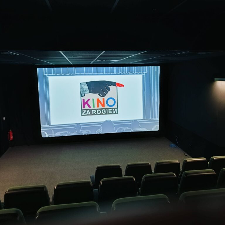 Małe, puste kino z zielonymi siedzeniami zwrócone jest w stronę dużego ekranu wyświetlającego logo, które głosi Kino za Rogiem z wskazującą ręką nad tekstem. Pomieszczenie jest słabo oświetlone, a ekran jest głównym źródłem światła.