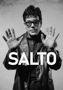 Czarno-białe zdjęcie mężczyzny w okularach przeciwsłonecznych i skórzanej kurtce trzymającego ręce w górze, z napisem „SALTO” na całej długości obrazu wydrukowanym pogrubionymi, białymi literami.
