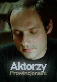 Mężczyzna z krótkimi, ciemnymi włosami patrzy w dół z lekkim uśmiechem. Ma na sobie czarną koszulę. Na dole obrazu widnieje tekst Aktorzy Prowincjonalni.