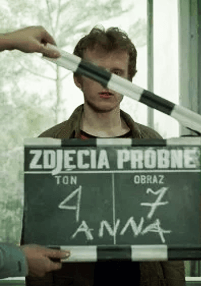Osoba stoi za klapsem filmowym z napisem „ZDJĘCIA PRÓBNE”, na którym widnieje ręcznie napisany tekst i widoczne imię „ANNA”, podczas gdy ktoś przesuwa klaps przed jej twarzą.