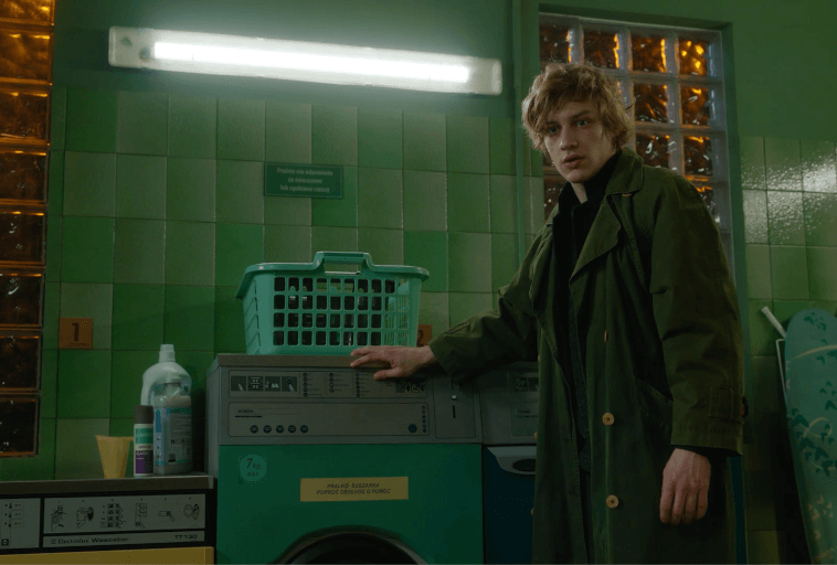 Młody mężczyzna z potarganymi blond włosami, ubrany w długi zielony płaszcz, stoi w pralni obok pralki z koszem na pranie na górze. W pomieszczeniu są zielone kafelki na ścianach i oświetlenie fluorescencyjne.