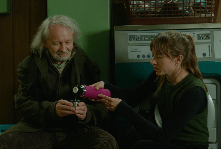 Starszy mężczyzna z siwymi włosami i brodą siedzi obok młodszej kobiety w pralni samoobsługowej. Kobieta podaje mu różowy termos, gdy siedzą razem przed pralką.