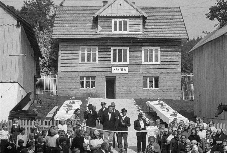 Tłum ludzi, w tym dzieci i dorośli, zbiera się przed drewnianym budynkiem z napisem SZKOŁA (szkoła). Na zewnątrz rozstawione są stoły z obrusami, a atmosfera wydaje się świąteczna.
