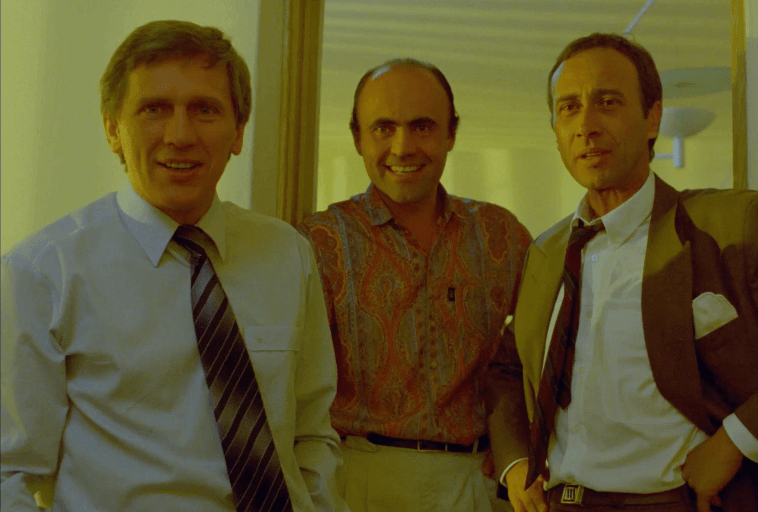 Trzech mężczyzn w strojach biznesowych stoi w pomieszczeniu, uśmiechając się do kamery. Dwóch ma na sobie garnitury i krawaty, a trzeci koszulę w paisley. Wyglądają, jakby byli w biurze lub na korytarzu z ciepłym oświetleniem.