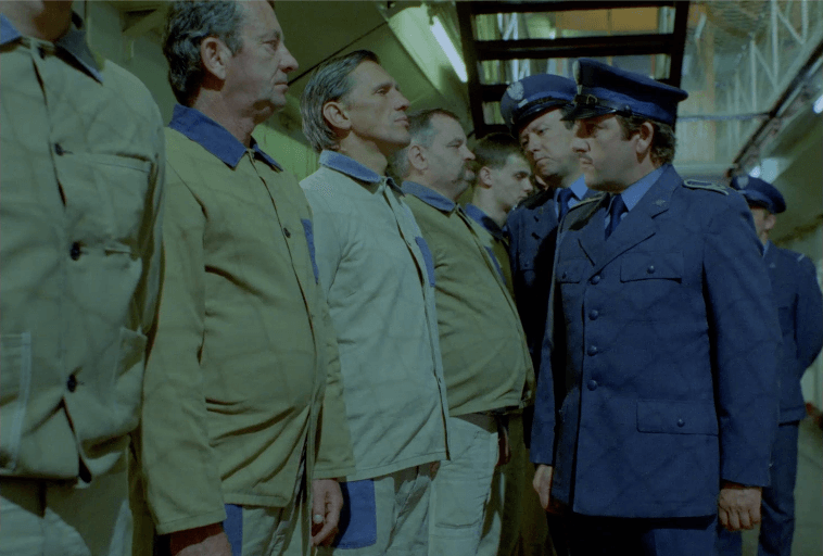 Grupa więźniów w beżowych mundurach stoi w kolejce naprzeciwko kilku surowo wyglądających strażników w niebieskich mundurach wewnątrz korytarza więziennego. Sceneria wydaje się napięta i poważna.