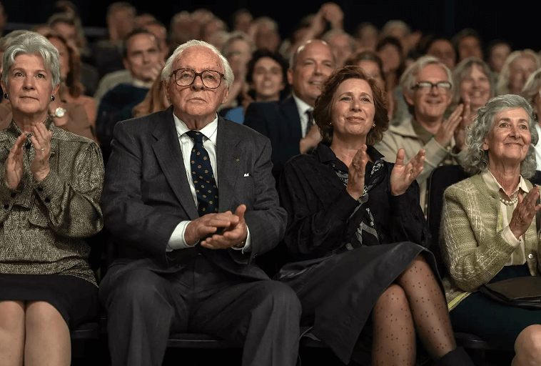 Grupa starszych dorosłych, ubranych formalnie, siedzi w pierwszym rzędzie audytorium. Niektórzy się uśmiechają i klaszczą, podczas gdy inni wyglądają na zamyślonych. Więcej osób wypełnia rzędy za nimi, wszyscy wydają się być zaangażowani w wydarzenie.