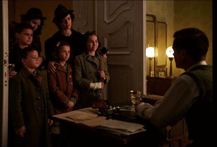 Pięcioro dzieci i dwoje dorosłych stoi razem w ciepło oświetlonym pokoju, naprzeciwko mężczyzny siedzącego przy biurku pokrytym papierami i maszyną do pisania. Sceneria wydaje się być zabytkowa, wszyscy ubrani są w staromodne ubrania.