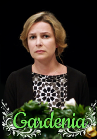 Kobieta o blond włosach, ubrana w czarno-biały, wzorzysty top i czarny kardigan, wygląda poważnie, stojąc za zielonymi liśćmi, na których u dołu widnieje ozdobny, zielony napis Gardenia.