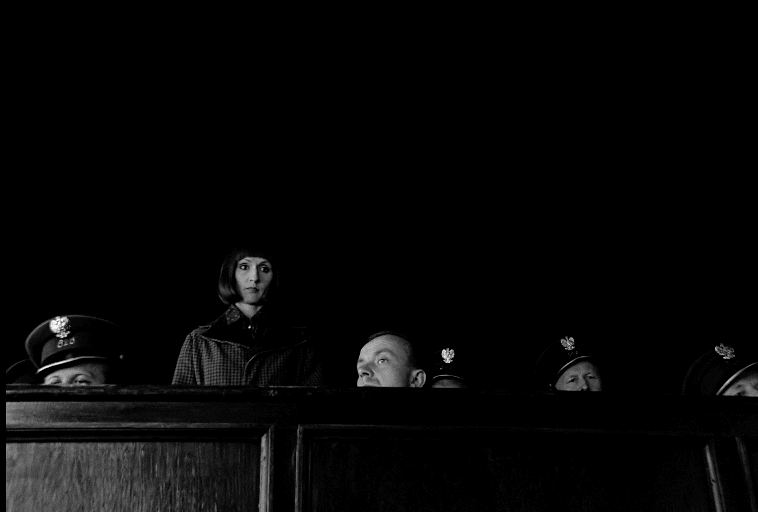 Kobieta wyróżnia się spośród kilku umundurowanych funkcjonariuszy, których wszyscy są częściowo widoczni za wysoką drewnianą barierą na surowym, czarnym tle.