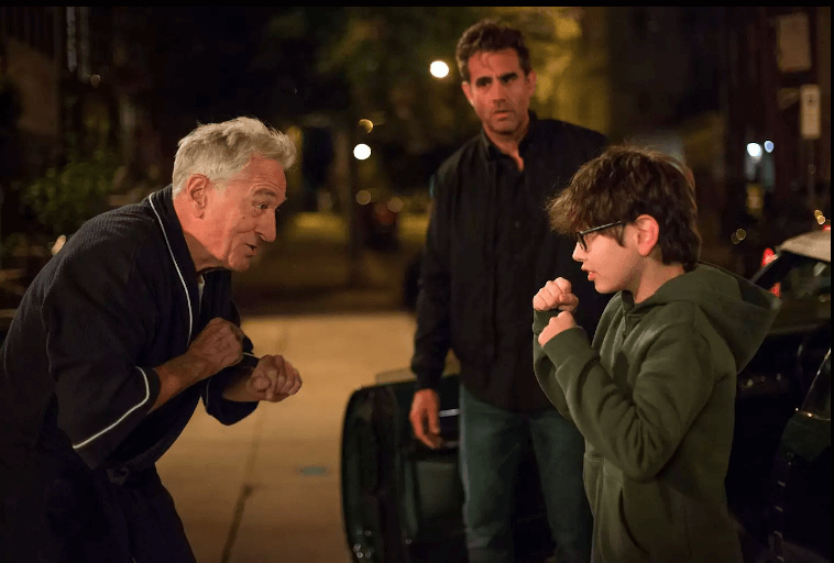 Starszy mężczyzna i chłopiec w okularach stoją naprzeciw siebie w zabawnych pozycjach bokserskich na ulicy nocą, podczas gdy zaniepokojony mężczyzna stoi między nimi i obserwuje scenę.