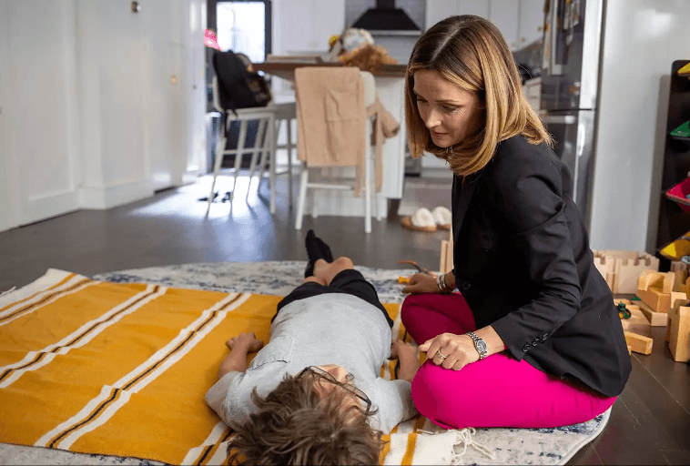 Kobieta klęczy obok dziecka leżącego na plecach na pasiastym żółto-białym kocu w domowym otoczeniu, oferując pocieszenie lub wsparcie. W tle widoczne są drewniane zabawki i meble kuchenne.