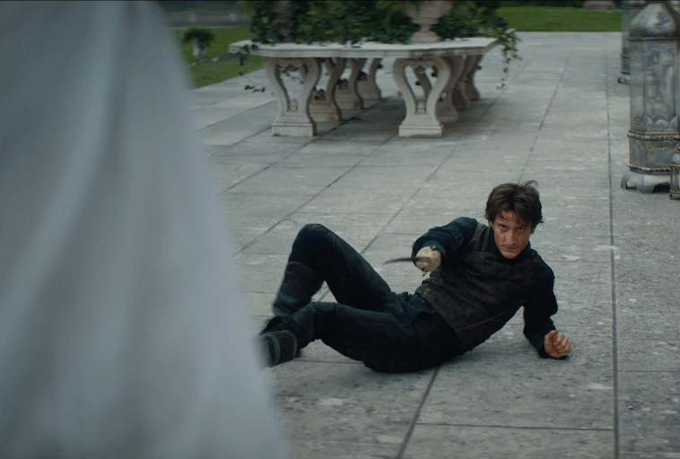 Mężczyzna w ciemnym ubraniu leży na ziemi, trzymając małe ostrze, wygląda na czujnego i napiętego. Ozdobne ławki i zieleń są widoczne w tle na wybrukowanym kamieniem terenie.