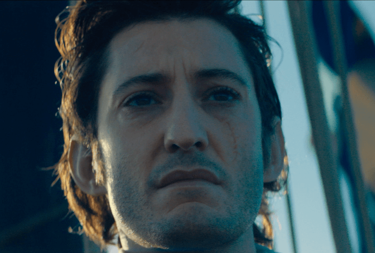 Zbliżenie mężczyzny z potarganymi, ciemnymi włosami i poważną miną, z widoczną blizną na prawym policzku, na rozmytym tle otoczenia.