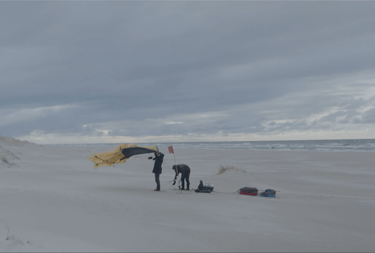 Dwie osoby stoją na wietrznej, pochmurnej plaży z wydmami, rozstawiając sprzęt. Jedna trzyma żółto-czarną tkaninę powiewającą na wietrze; sprzęt i torby leżą na piasku w pobliżu. Widać fale oceanu.