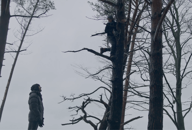 Osoba w zimowym płaszczu stoi na ziemi i patrzy na inną osobę wspinającą się na wysokie, bezlistne drzewo na tle zachmurzonego nieba.