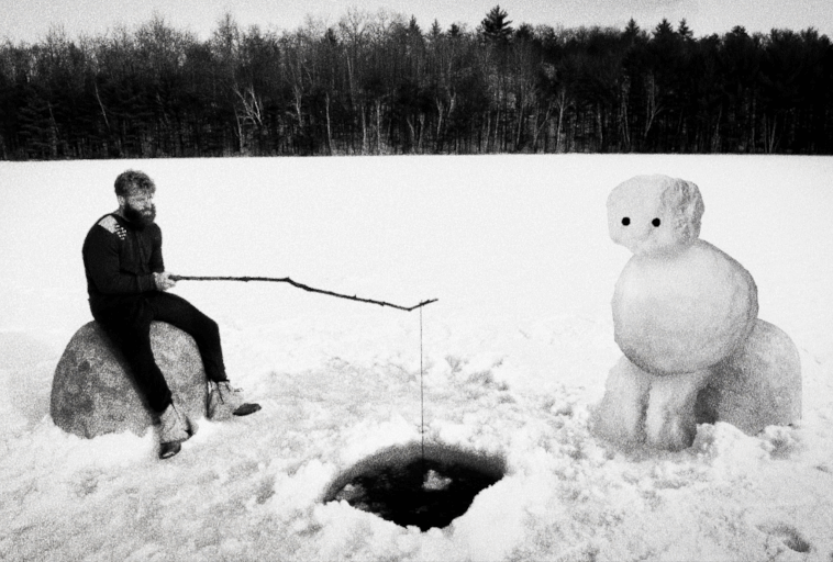 Osoba siedzi na skale i łowi ryby przez dziurę w lodzie zaśnieżonego jeziora, patrząc na dużego bałwana z ciemnymi oczami; drzewa wyznaczają tło. Scena jest czarno-biała.