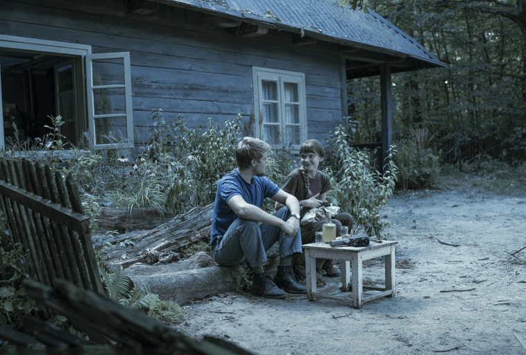 Dwie osoby siedzą na kłodzie przed rustykalnym drewnianym domem, rozmawiają i się uśmiechają. Przed nimi znajduje się mały stolik z jedzeniem i napojami, a scena jest otoczona zielenią i drzewami.