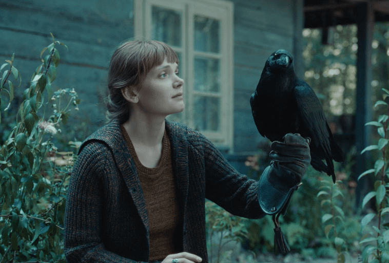 Kobieta z krótkimi włosami w swetrze i rękawiczkach siedzi na zewnątrz, spokojnie trzymając czarnego kruka na dłoni w rękawiczce. Otaczają ją zielone rośliny, a w tle widać drewniany dom z oknami.