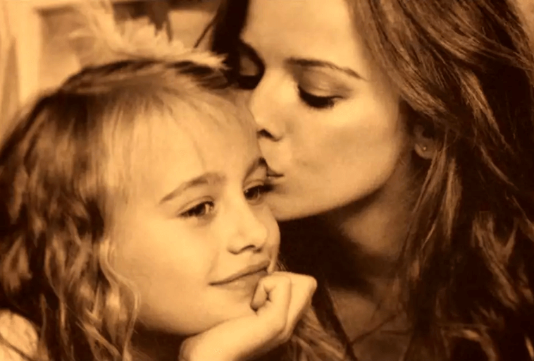 Kobieta delikatnie całuje młodą dziewczynę w skroń. Dziewczyna uśmiecha się delikatnie, opierając brodę na dłoni. Obraz jest ciepło oświetlony, przekazując poczucie czułości i uczucia między nimi.