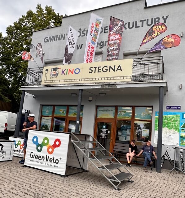 Przód kina Stegna z flagami plakatowymi z filmami Marvela i Gwiezdnych Wojen. Trzy osoby siedzą i stoją przy rampie wejściowej. Na barierce na zewnątrz zamontowany jest znak Green Velo.
