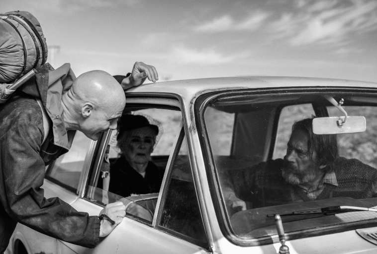 Łysy mężczyzna z plecakiem pochyla się do otwartego okna samochodu, rozmawiając ze starszą kobietą na siedzeniu pasażera i starszym mężczyzną na siedzeniu kierowcy. Scena jest na zewnątrz w czerni i bieli.