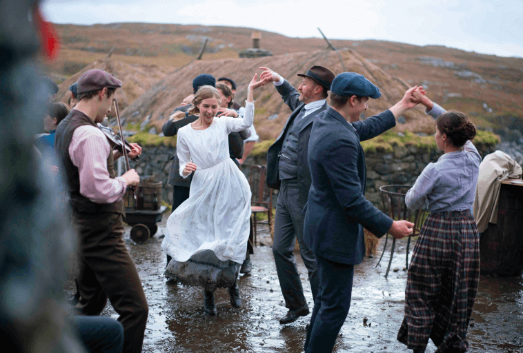 Grupa ludzi ubranych w staromodne stroje tańczy radośnie na zewnątrz, na błotnistym gruncie, podczas gdy mężczyzna gra na skrzypcach. W tle widać wzgórza i rustykalne budynki.