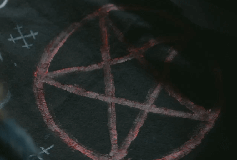 Czerwony pentagram jest narysowany na ciemnej powierzchni z dodatkowymi białymi symbolami kredowymi w pobliżu. Obraz jest słabo oświetlony, co tworzy tajemniczą atmosferę.