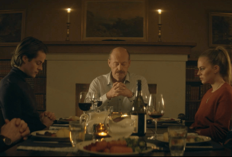 Trzy osoby siedzą uroczyście przy stole z winem i jedzeniem, z dłońmi złożonymi lub splecionymi, jakby modliły się lub rozmyślały. Pokój jest ciepło oświetlony świecami, a w tle widać kominek.