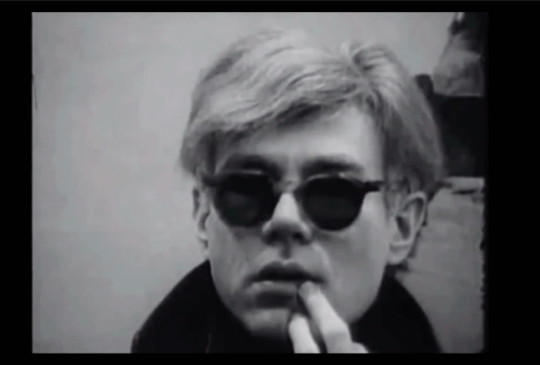 Czarno-białe zdjęcie przedstawiające osobę o jasnych włosach, w ciemnych okularach przeciwsłonecznych, dotykającą ust i patrzącą przed siebie z neutralnym wyrazem twarzy.