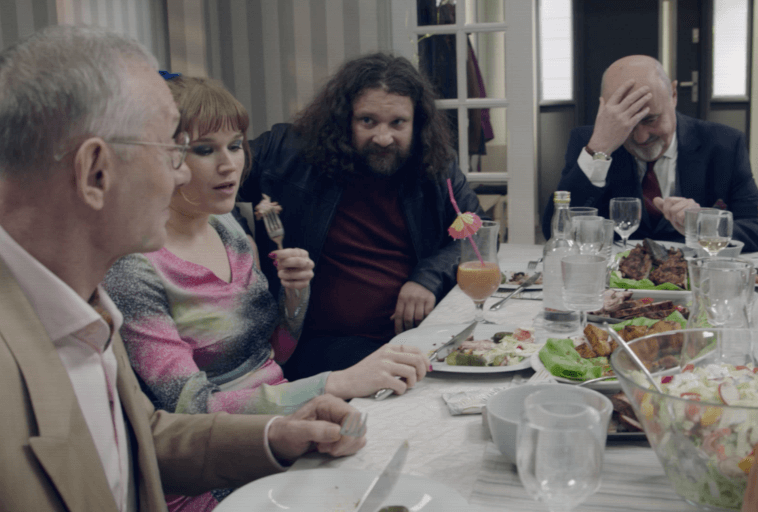 Cztery osoby siedzą przy stole jadalnym z talerzami pełnymi jedzenia i napojów. Dwóch mężczyzn nosi garnitury, jedna kobieta w kolorowej sukience je, a drugi mężczyzna z długimi włosami patrzy na nią. Jeden mężczyzna trzyma się za głowę z frustracją.