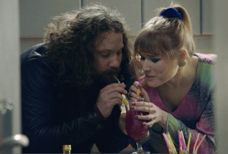 Mężczyzna i kobieta piją jasnoróżowy napój przez słomki, pochylając się blisko i uśmiechając się. Kobieta ma na sobie kolorowy strój i niebieską gumkę do włosów; mężczyzna ma długie kręcone włosy i brodę.