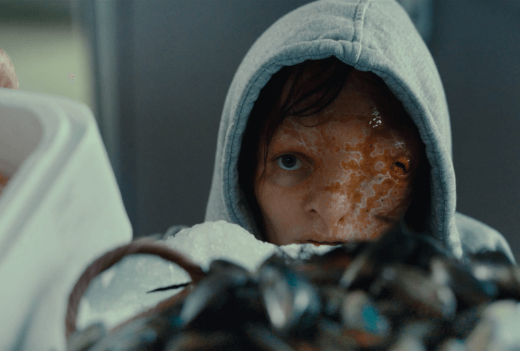 Osoba ubrana w szary kaptur spogląda w górę, z jedną stroną twarzy pokrytą łuskowatym, pomarańczowym nalotem. Jest częściowo otoczona bąbelkami i przedmiotami na pierwszym planie, co tworzy napiętą i tajemniczą atmosferę.