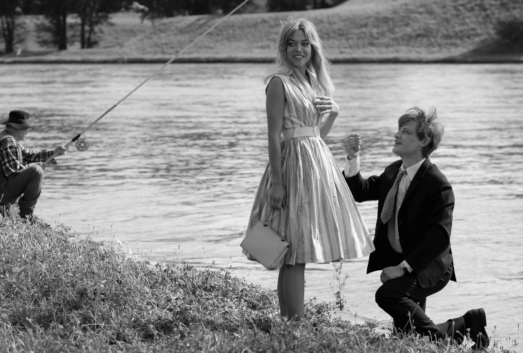 Mężczyzna w garniturze klęczy nad rzeką, trzymając pierścionek w stronę kobiety w sukience, pozornie się oświadczając. Kobieta wygląda na zaskoczoną. W tle ktoś łowi ryby na brzegu rzeki. Obraz jest czarno-biały.