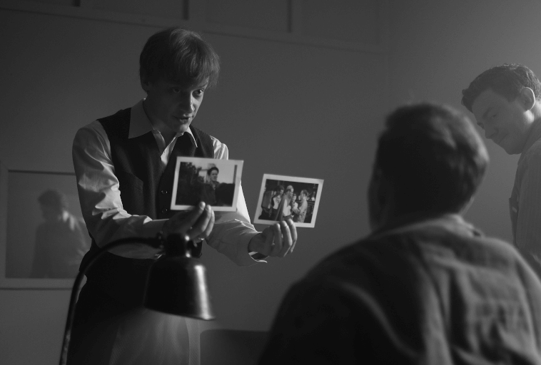 Osoba w kamizelce pokazuje dwa zdjęcia siedzącemu mężczyźnie w słabo oświetlonym pokoju, podczas gdy inna osoba patrzy. Scena jest czarno-biała, co tworzy dramatyczną, napiętą atmosferę.