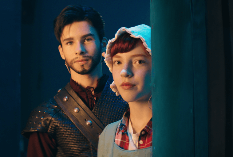 Mężczyzna w zbroi w stylu średniowiecznym i kobieta w czepcu i prostej sukience stoją razem, patrząc przed siebie. Niebieskie i zielone oświetlenie tworzy dramatyczną atmosferę.