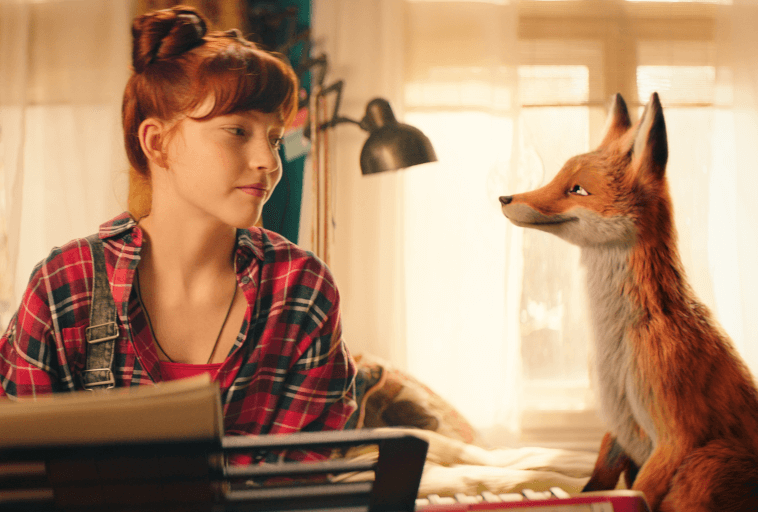 Młoda kobieta z rudymi włosami w kraciastej koszuli siedzi w środku, uśmiechając się do animowanego lisa obok niej. Wydaje się, że spędzają chwilę przy biurku, a łagodne światło słoneczne wpada przez okno.