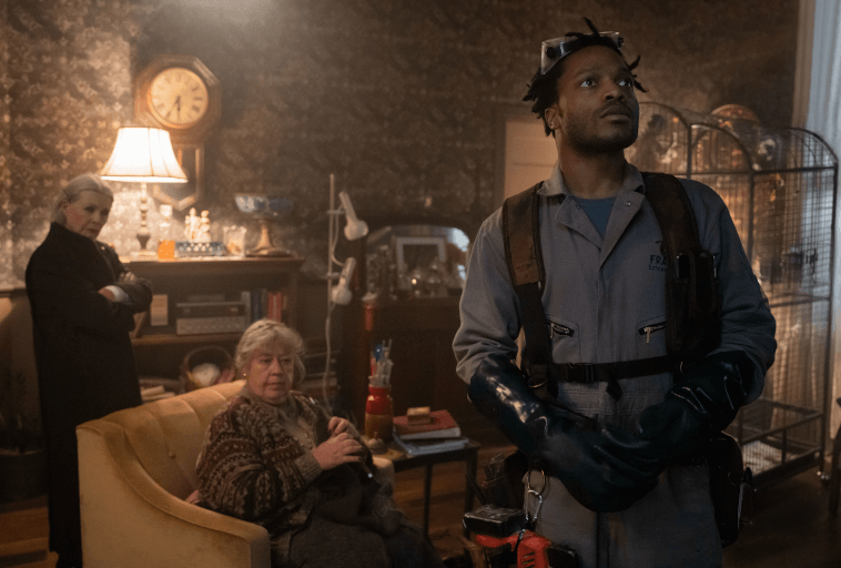 Mężczyzna w gumowych rękawiczkach i roboczym kombinezonie stoi w salonie, patrząc w górę. Dwie starsze kobiety, jedna siedzi na kanapie, a druga stoi ze skrzyżowanymi ramionami, obserwują go. Pokój jest zagracony, z zabytkowymi dekoracjami i klatką dla ptaków.