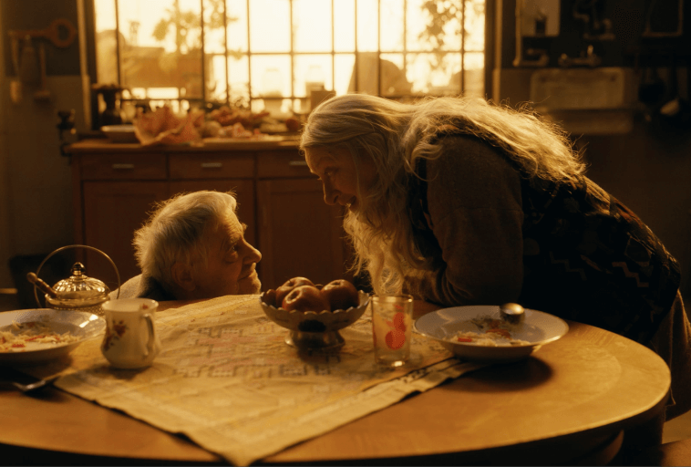 Starsza para uśmiecha się do siebie ciepło przy kuchennym stole nakrytym do śniadania, skąpanym w złotym świetle słonecznym z dużego okna za nimi. Atmosfera jest przytulna i intymna.