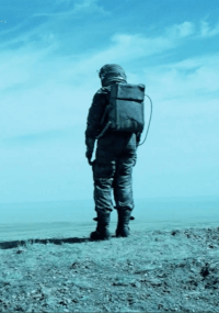 Osoba ubrana w skafander kosmiczny lub inny sprzęt ochronny stoi samotnie na jałowym terenie, zwrócona twarzą w stronę rozległego, pustego krajobrazu pod błękitnym niebem z delikatnymi chmurami.