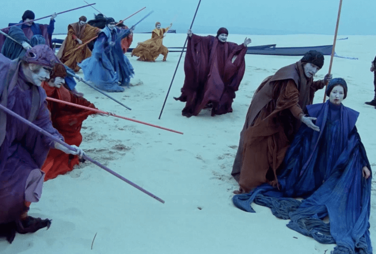 Grupa ludzi w powłóczystych czerwonych, niebieskich i fioletowych szatach z pomalowanymi twarzami stoi i klęczy na piaszczystej plaży, trzymając kolorowe kije. W tle widać morze i drewnianą konstrukcję.