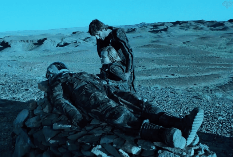 Dwie osoby klęczą obok mężczyzny w skafandrze kosmicznym leżącego na stosie kamieni na jałowym, błękitnym krajobrazie pustyni z odległymi wzgórzami pod czystym niebem.