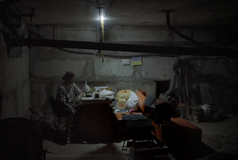 Osoba siedzi sama przy zagraconym biurku pod słabym światłem w ciemnej, niedokończonej piwnicy. Nad głową biegną odsłonięte rury, a różne przedmioty i gruz są porozrzucane po całym pomieszczeniu.